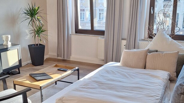 A modern bedroom with a large bed, a grey sofa, a bedside table, a potted plant, and a chandelier. - Stylish apartment in the Gründerzeit district (Chemnitz)