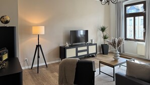 A modern living room with a flat-screen TV, a floor lamp, a black cabinet, and a large window with curtains. - Stylish apartment in the Gründerzeit district (Chemnitz)