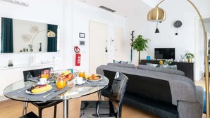 A modern living room with a grey sofa, a round glass table with breakfast, a TV, and a gold pendant light.