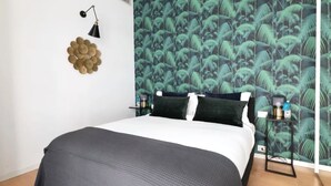 A bedroom with a bed, a wall-mounted lamp, a decorative wall hanging, and a nightstand with a lamp.