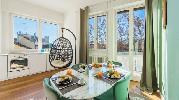 A modern kitchen with a hanging chair, a dining table set with breakfast, and large windows with a city view. - Sweett | Mentana IV - Two Bedroom Apartment, Sleeps 3 (Milano)