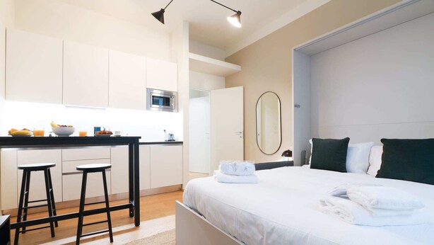 A modern hotel room with a bed, a small kitchen area, and a dining table.