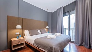 Moderni hotellihuone, jossa on suuri sänky, puiset yöpöydät ja parvekkeelta avautuva näkymä kaupunkiin.