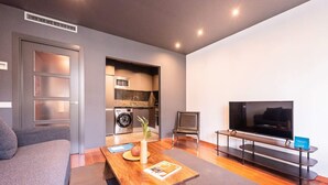 A modern living room with a sofa, a wooden coffee table, a television, and a built-in kitchen area with appliances. - Sweett | Roma 5B - One Bedroom Apartment, Sleeps 4 (Barcelona)