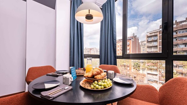 A dining area with a round table, red upholstered chairs, a breakfast spread including croissants, grapes, and orange juice, and a view of urban buildings through large windows with blue curtains. - Sweett | Roma 5B - One Bedroom Apartment, Sleeps 4 (Barcelona)