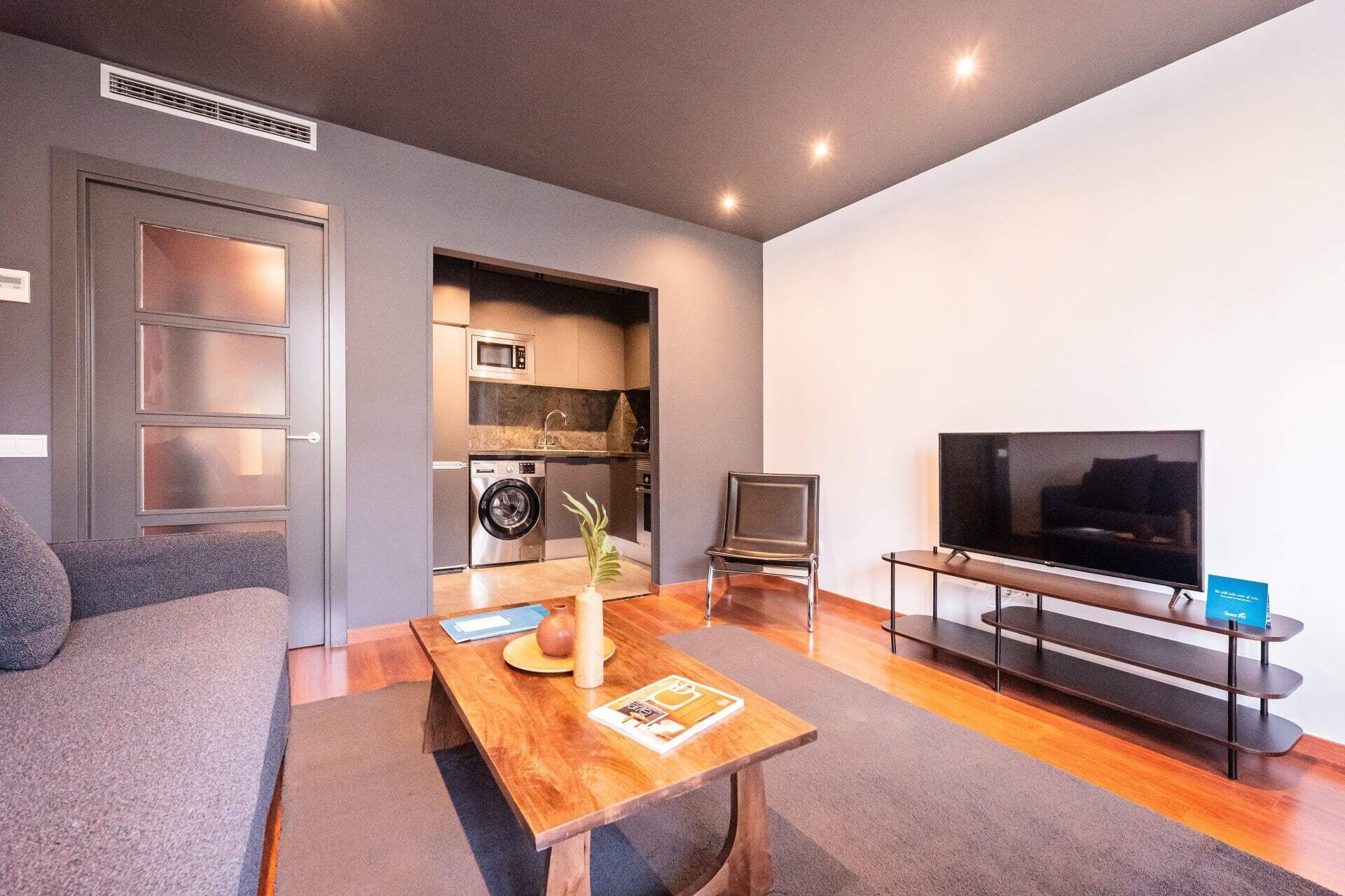 A modern living room with a sofa, a wooden coffee table, a television, and a built-in kitchen area with appliances.