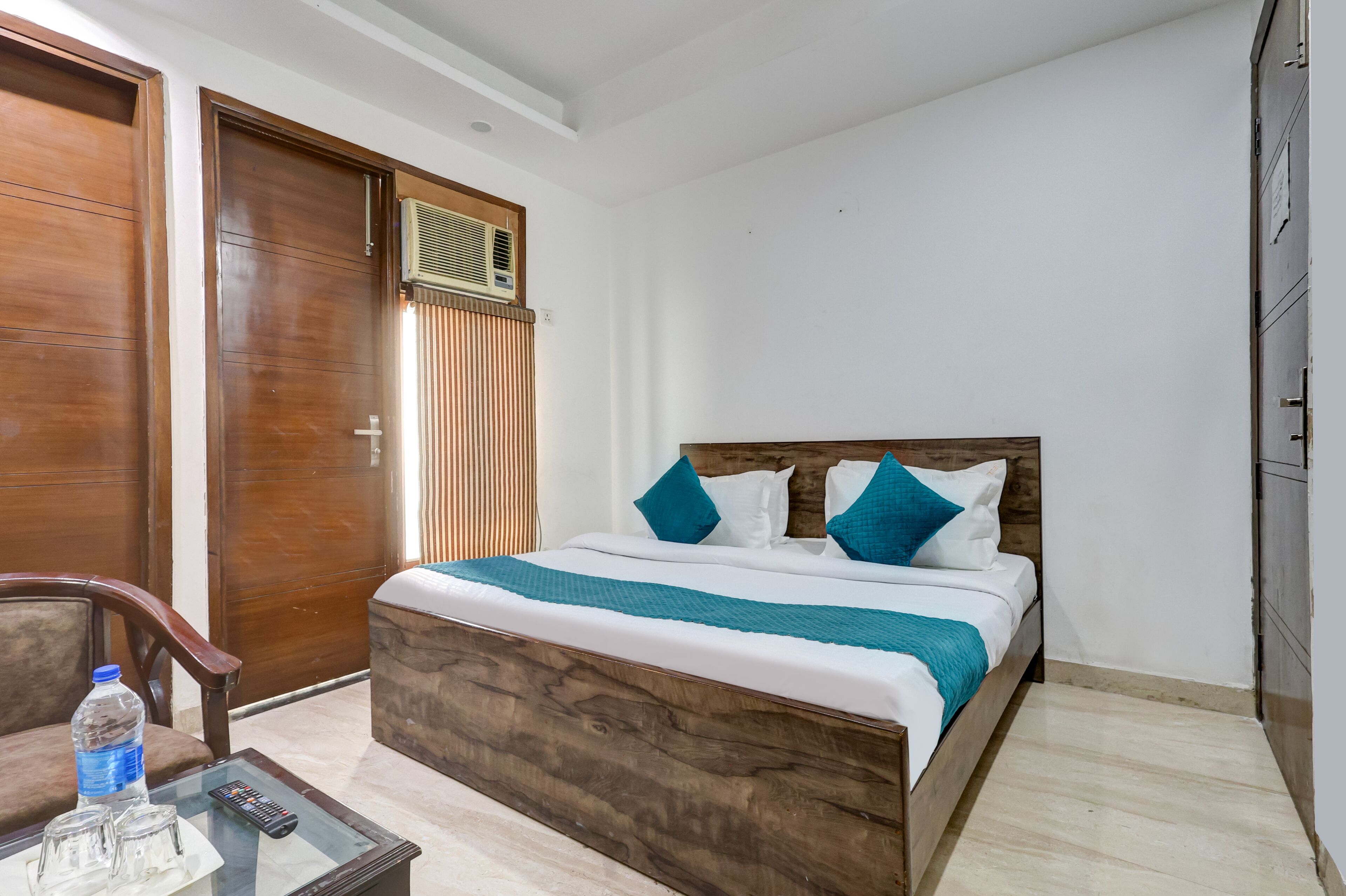 Habitación de hotel ordenada con una cama de madera, una mesita de vidrio con un control remoto y una botella de agua.