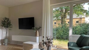 Un salón moderno con un sofá blanco, un sillón verde, un televisor de pantalla plana montado en la pared y un amplio ventanal con vistas a un jardín y edificios.