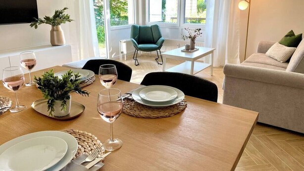 Un comedor con una mesa de madera dispuesta para dos personas, un sillón verde, un sofá blanco y una lámpara de pie.