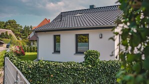 A white house with a tiled roof, two windows, and a hedge in front.