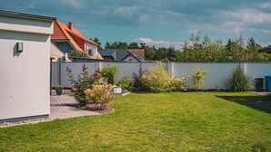 A residential area with a well-maintained lawn, a white building with a red roof, and a variety of flowering plants.