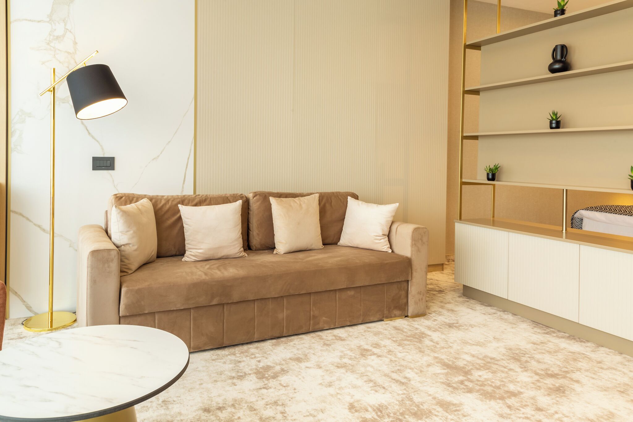 A modern living room with a beige sofa, a round coffee table, a floor lamp, and a shelving unit with decorative items.