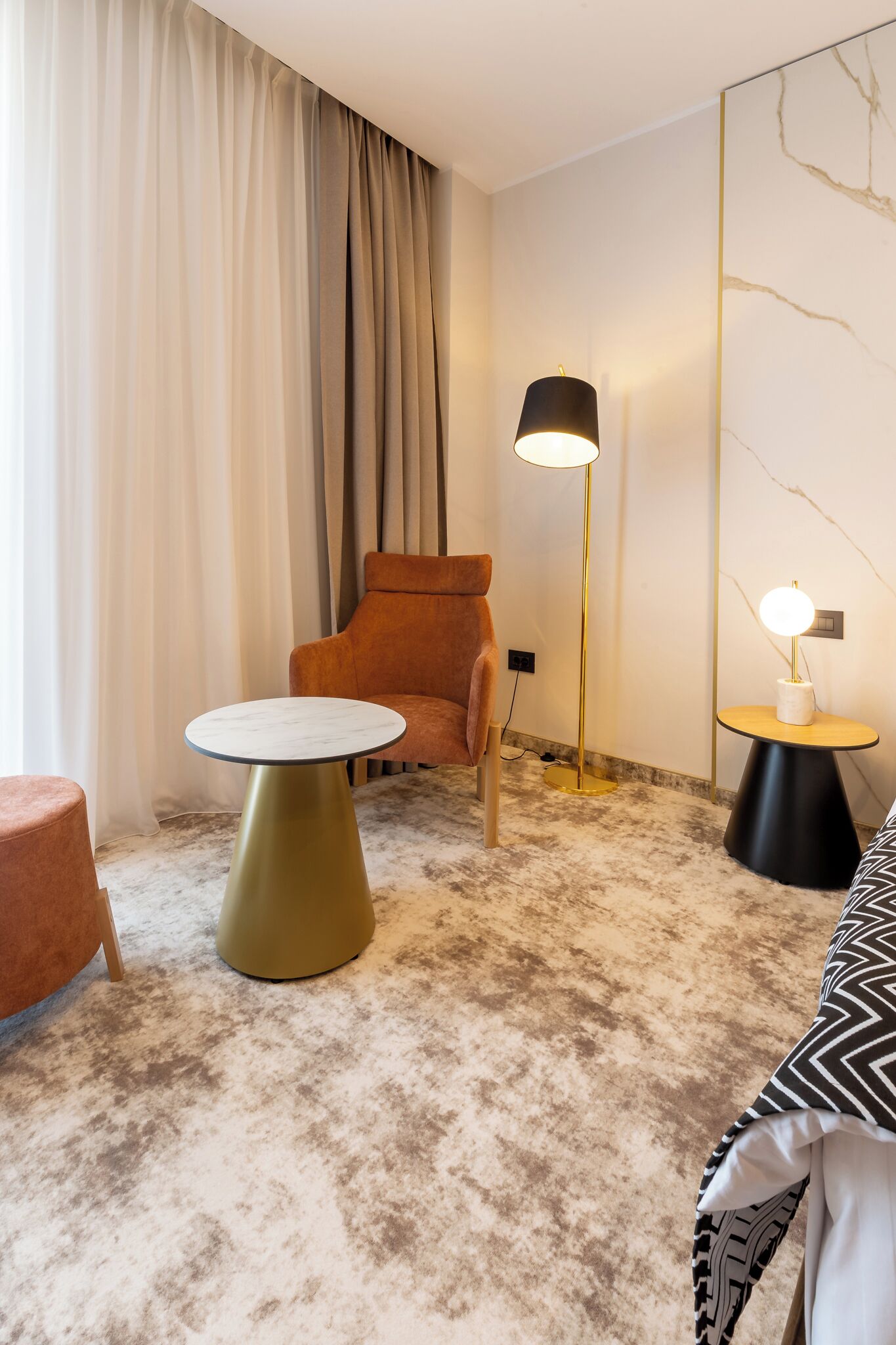 A modern hotel room with a round table, a plush orange chair, a floor lamp, and a marble-patterned wall.