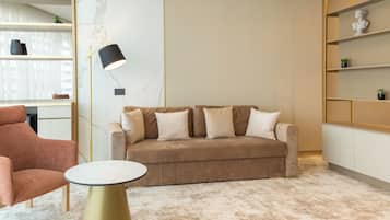 Een moderne woonkamer met een beige bank, een ronde tafel en een roze fauteuil. Er is een boekenkast met decoratieve objecten en een vloerlamp.