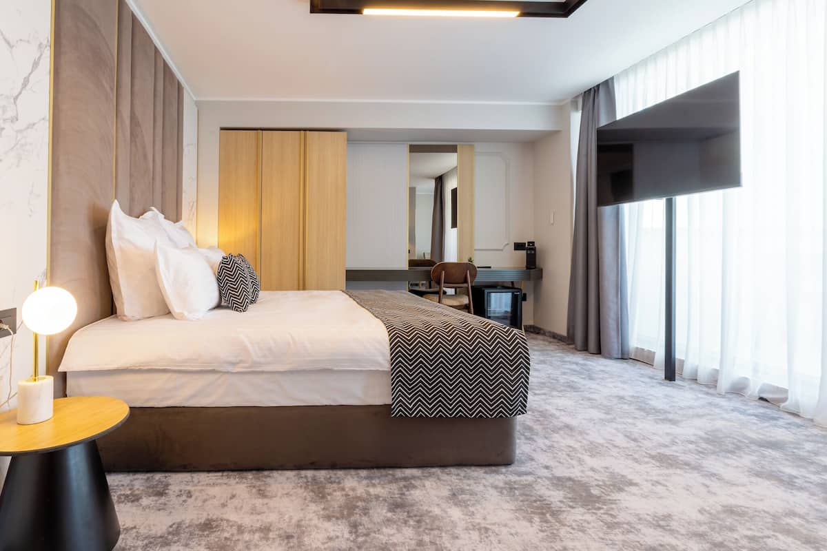 Ein modernes Hotelzimmer mit einem großen Bett, einem Nachttisch mit Lampe, einem Schreibtisch mit Stuhl und einem großen Fenster mit Vorhängen.