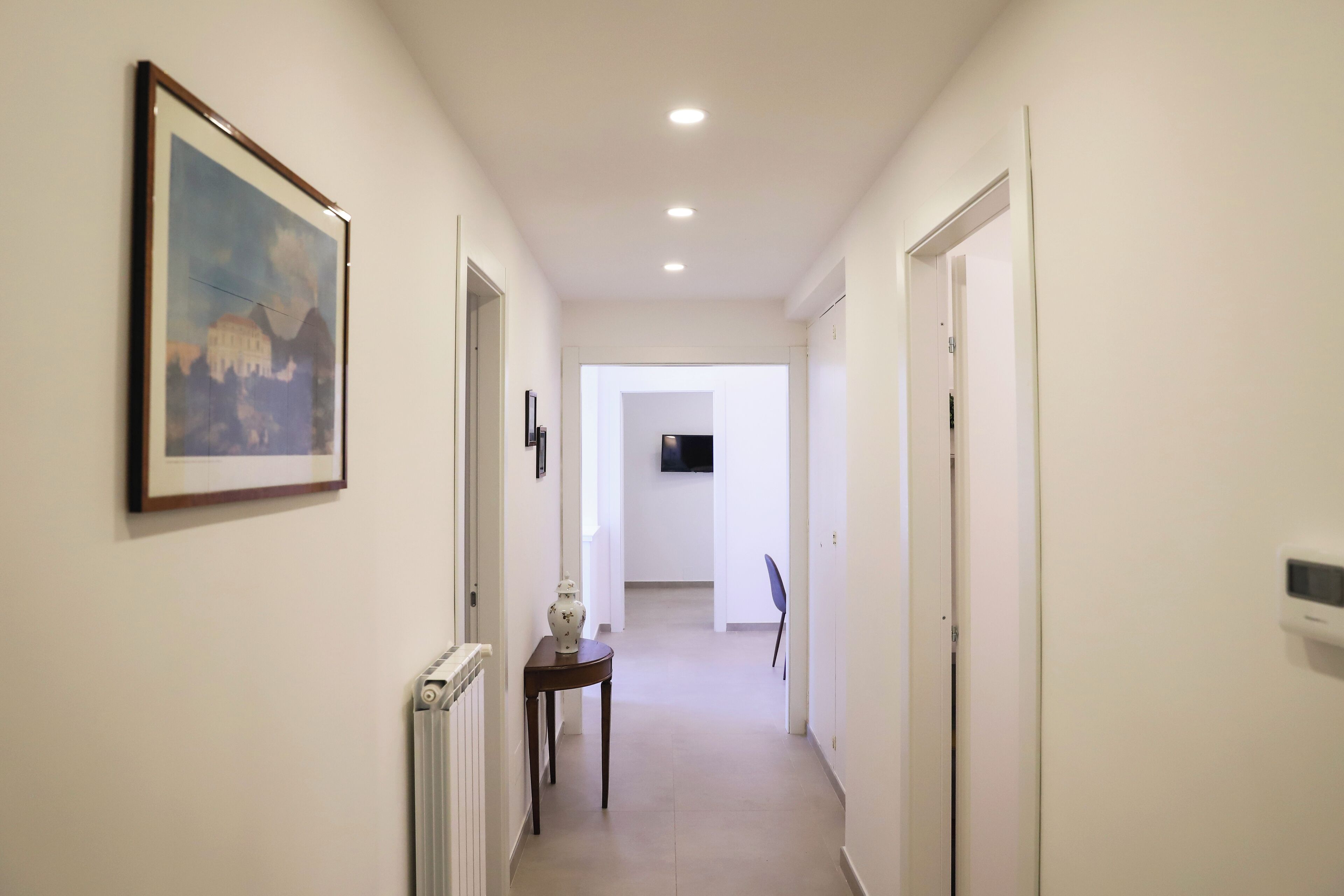 A hallway with a framed picture on the left, a radiator, and a small table with a vase. The hallway leads to a room with a TV.