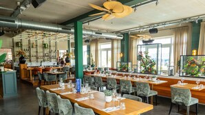 Un restaurant spacieux, doté de nombreuses tables et chaises, de grandes fenêtres avec des rideaux et de ventilateurs de plafond.