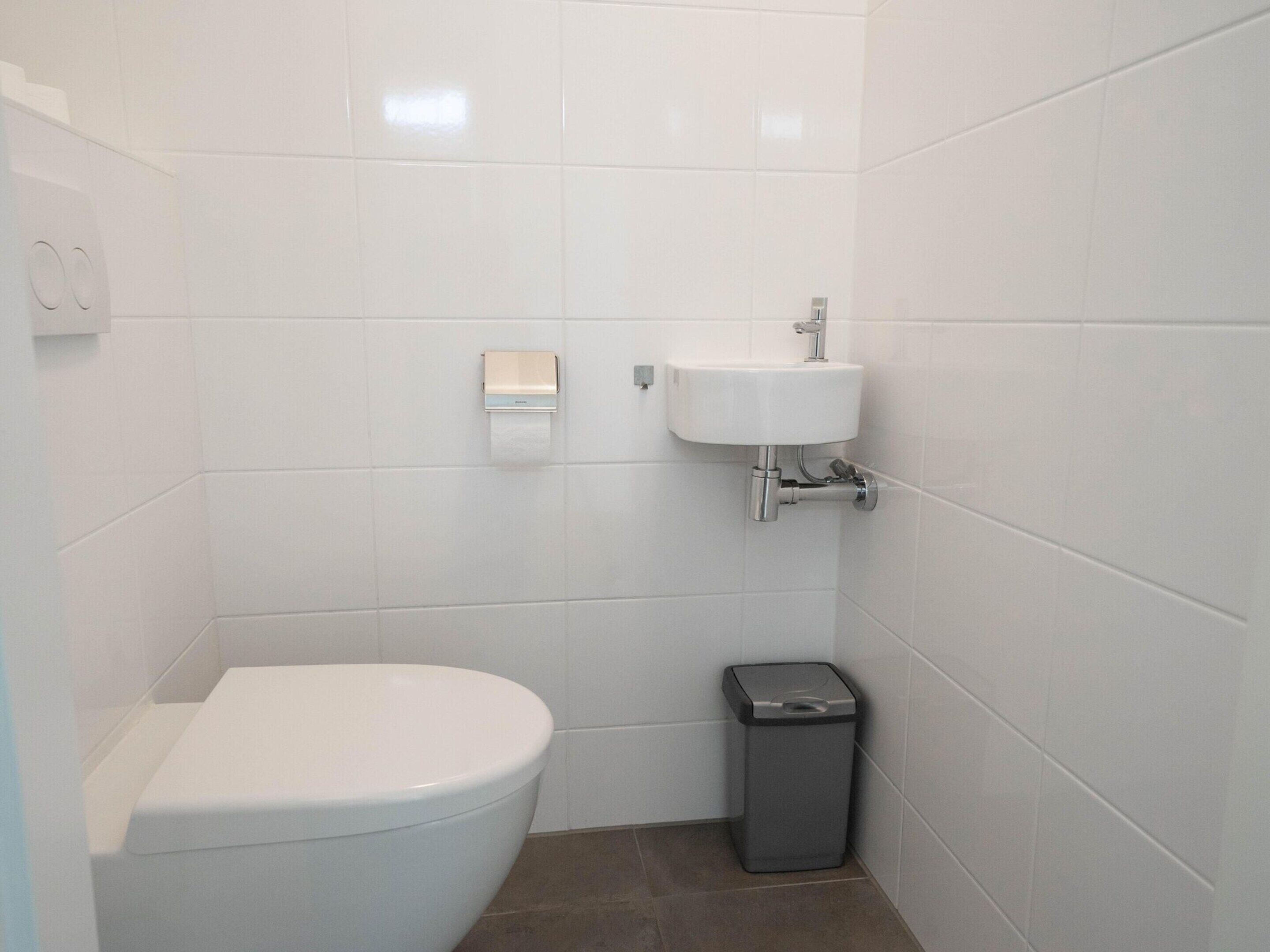 Een kleine, schone badkamer met witte tegels, een toilet, een wastafel en een prullenbak.