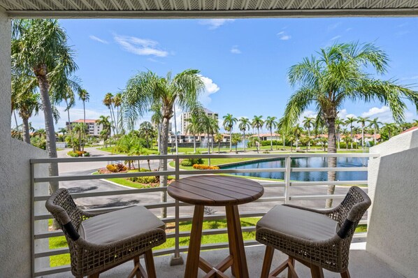 A balcony with wicker chairs and a table, overlooking a pool and palm trees.