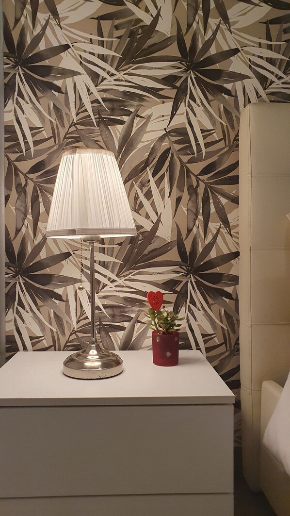A bedside table with a lamp and a red flower pot against a wall with a leaf pattern.