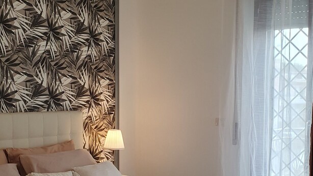 Ein modernes Schlafzimmer mit einem großen Bett, einer Zierwandverkleidung, einer hängenden Leuchte und Blick durch transparente Vorhänge.