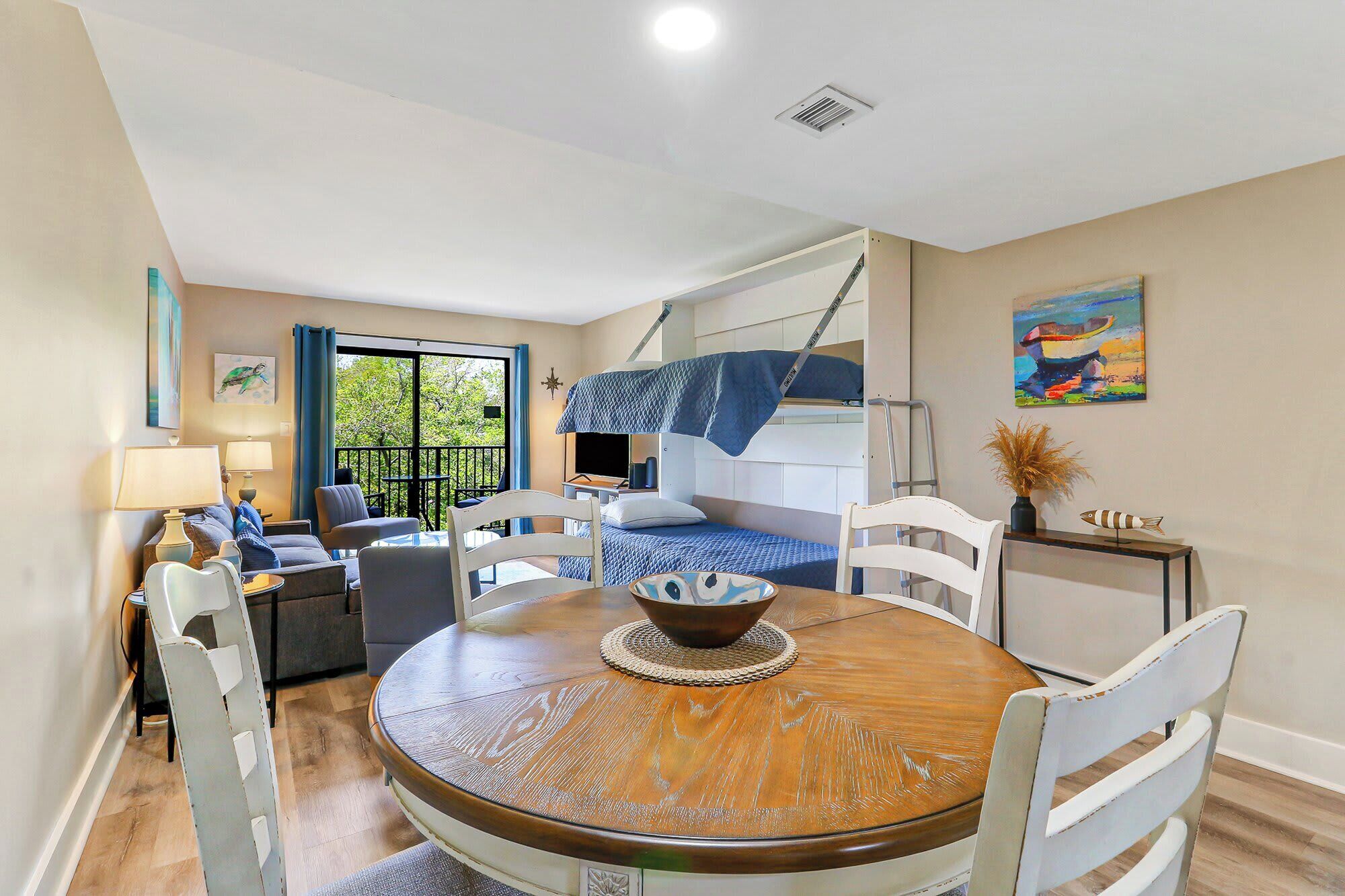 A room with a bunk bed, a dining table with chairs, a sofa, and a balcony with a view of trees.