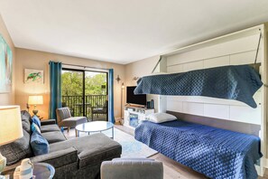 Habitación compacta con cama, sofá, una mesita y balcón con vista a árboles.