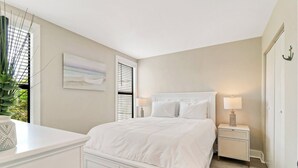 Une chambre à coucher comprenant un lit, des tables de chevet, une commode, une lampe, une fenêtre avec des stores et une plante.