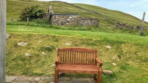 Un banc en bois se trouve dans un champ herbeux, avec un mur de pierres et un bâtiment en pierre en arrière-plan.