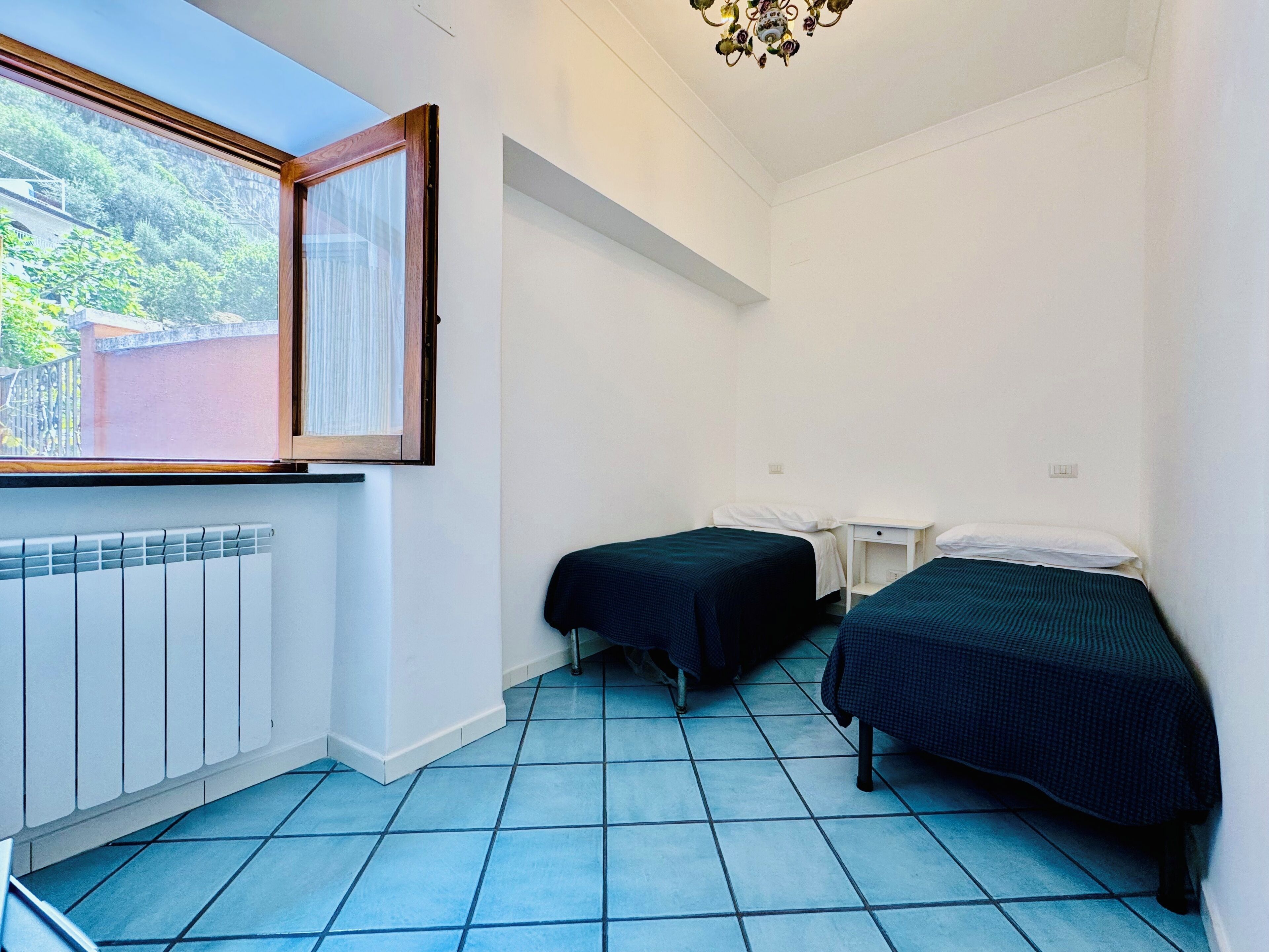 A small, clean room with two single beds, a window with a view of greenery, a radiator, and a white wall.