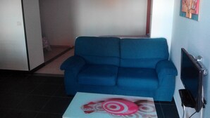 Un salon avec un canapé bleu, une petite table blanche ornée d’un objet décoratif, un téléviseur à écran plat et un tableau accroché au mur.