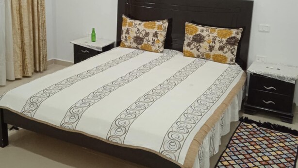 Une chambre à coucher avec un lit, deux tables de chevet, une bouteille verte, un tapis à motifs et une fenêtre avec des rideaux.