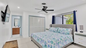 Une chambre à coucher comprenant un lit, un ventilateur de plafond, une table de chevet, un téléviseur, une armoire et une fenêtre avec des rideaux.