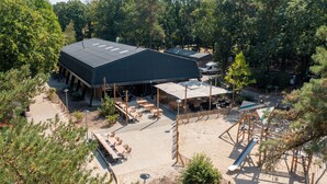 A large building with a flat roof, surrounded by trees and a sandy area with outdoor seating. - Bungalow in Park De Peel - Car-free Retreat (Vlierden)