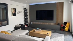 Ein modernes Wohnzimmer mit einem großen Flachbildfernseher an der Wand, einem Holztisch, grauen Sofas, einem Schuhregal und einer kleinen Sitzecke mit Kissen.