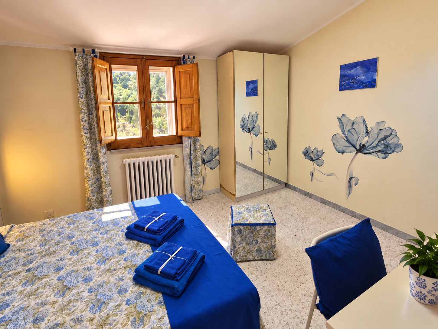 Una habitación con cama, armario, radiador, ventana con cortinas y un cuadro floral en la pared.
