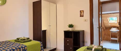 Quarto com duas camas, um guarda-roupa, um pequeno armário e uma porta que dá para uma varanda.