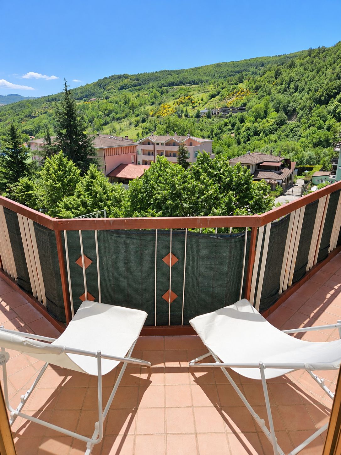 A balcony with two white chairs, a view of a green hillside, and buildings in the background.