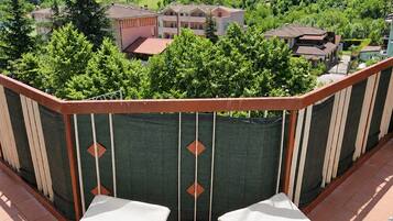 A balcony with two white chairs, a view of a green hillside, and buildings in the background.