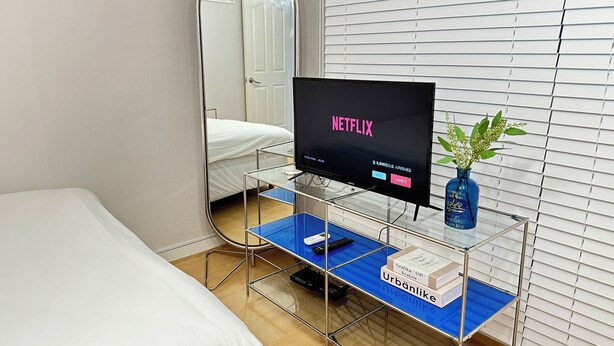 Hotellihuone, jossa on sänky, jalustalla oleva televisio, peili ja ikkuna, jossa on sälekaihtimet.