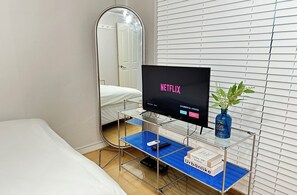 A hotel room with a bed, a TV on a stand, a mirror, and a window with blinds.