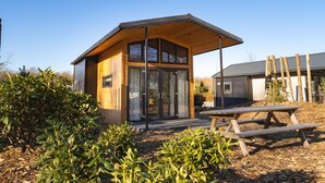 Une cabane moderne en bois, dotée d’un porche couvert, de grandes fenêtres et d’une table de pique-nique, située dans un espace aménagé.