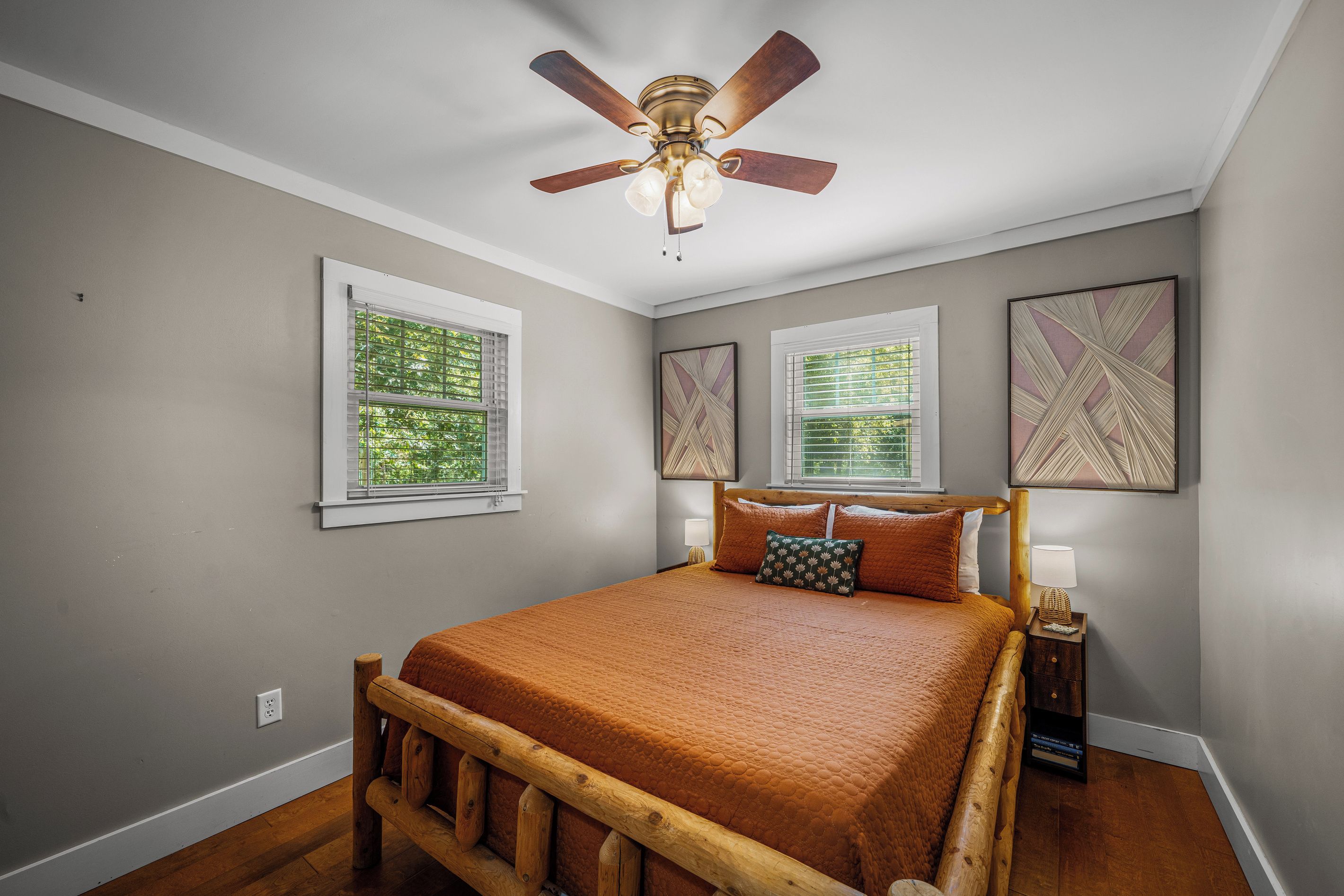 A bedroom with a bed, wooden headboard, a ceiling fan, two windows with blinds, and two wall-mounted artworks.