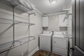 Une buanderie équipée de deux machines à laver et deux sèche-linge superposés, d’étagères métalliques et d’une horloge murale.