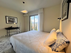 A bedroom with a bed, a desk, a chair, a hanging light fixture, and a window with curtains. - Terrasse des Agaves (Niza)
