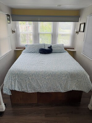 Un dormitorio con una cama, dos ventanas con persianas, un repuesto a cuadros y un cojín decorativo con forma de ballena.