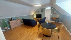 Un espacio habitable tipo loft con techo alto, piso de madera, un sofá azul oscuro, una mesa de comedor redonda con sillas, un televisor y un pequeño área de trabajo con escritorio y silla.