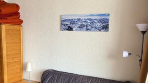Ein Zimmer mit grauem Sofa, einem Holzschrank, einem kleinen Nachttisch mit Lampe und einem Wandbild von Bergen.