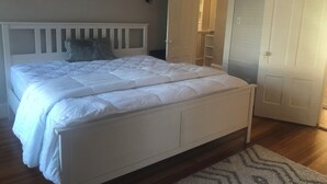Ein Schlafzimmer mit einem weißen Bett, einem Holz-Kopfteil, einem grauen Kopfkissen, einer weißen Wand, einer weißen Tür und einem weißen Nachttisch.