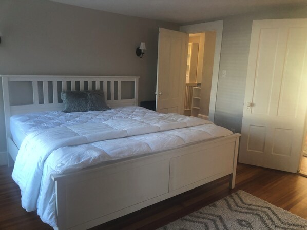 Quarto com cama branca, cabeceira de madeira, travesseiro cinza, parede branca, porta branca e mesa de cabeceira branca.